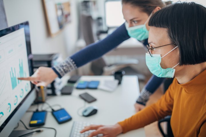 Two women, working together on computer in office with protective masks, to protect themselves against coronavirus.