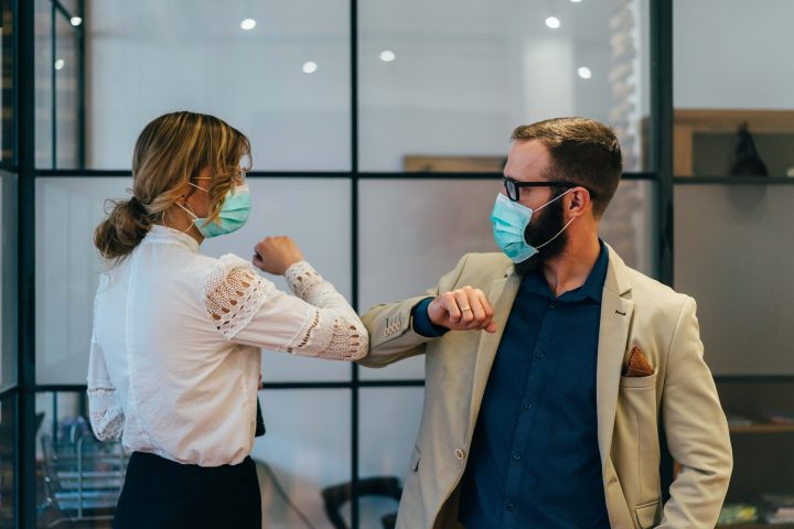 Eine Frau und ein Mann begrüßen einander, indem sie ihre Ellenbogen aneinander bringen. Beide tragen FFP2 Masken. Der Hintergrund zeigt Büroräumlichkeiten.
